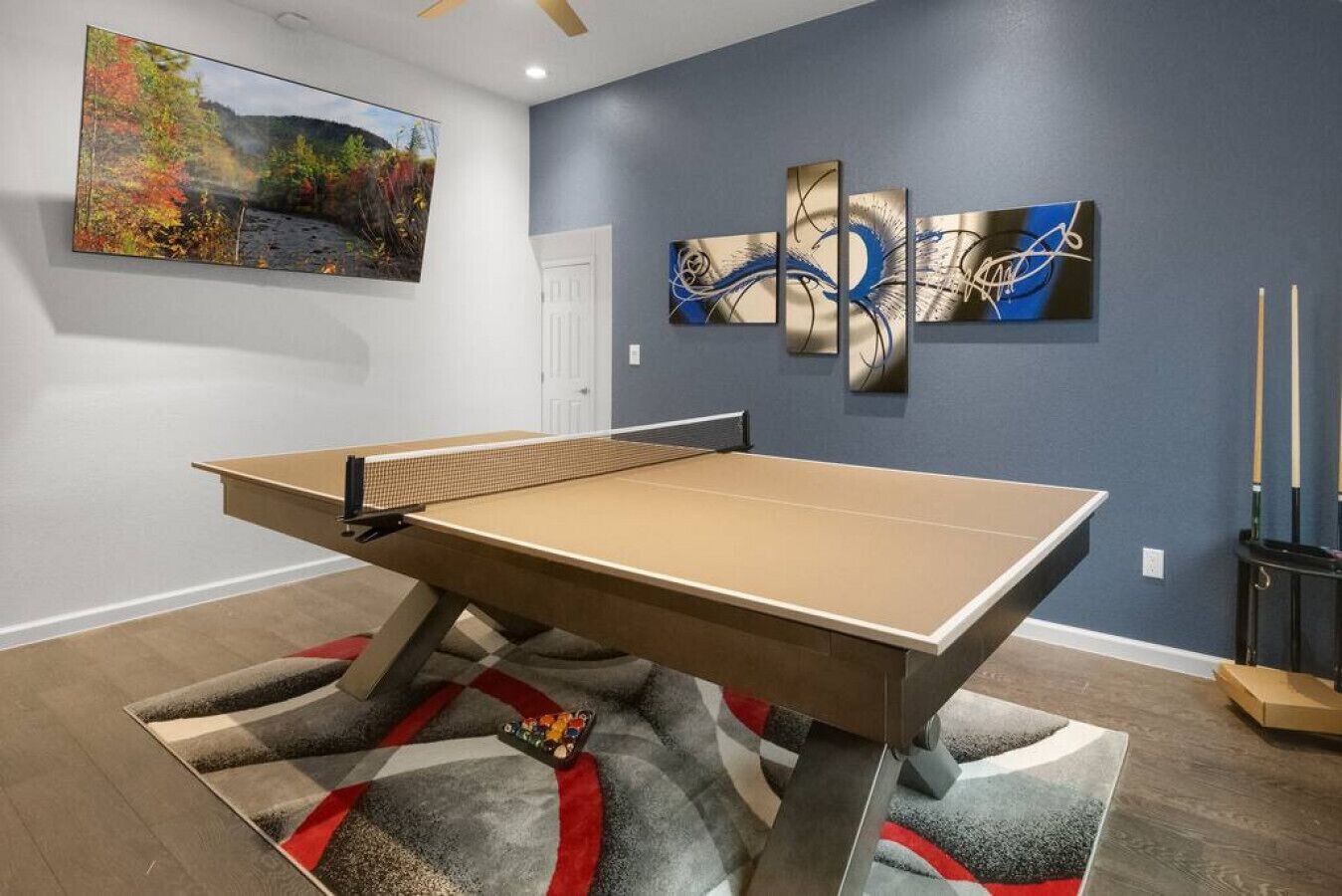 Une table de ping-pong dans une pièce dotée d’un tapis à carreaux, d’œuvres d’art murales abstraites et d’un tableau représentant un paysage.