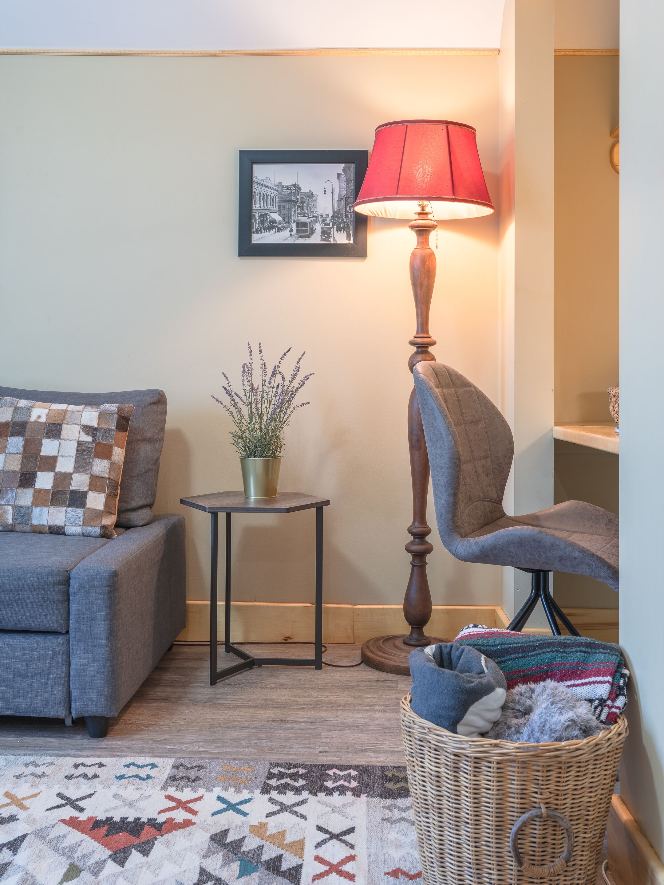 Ein Wohnzimmer mit grauem Sofa, einer roten Lampe, einem gerahmten Bild an der Wand, einem kleinen Tisch mit Pflanze und einem Korbkörbchen mit Decken.