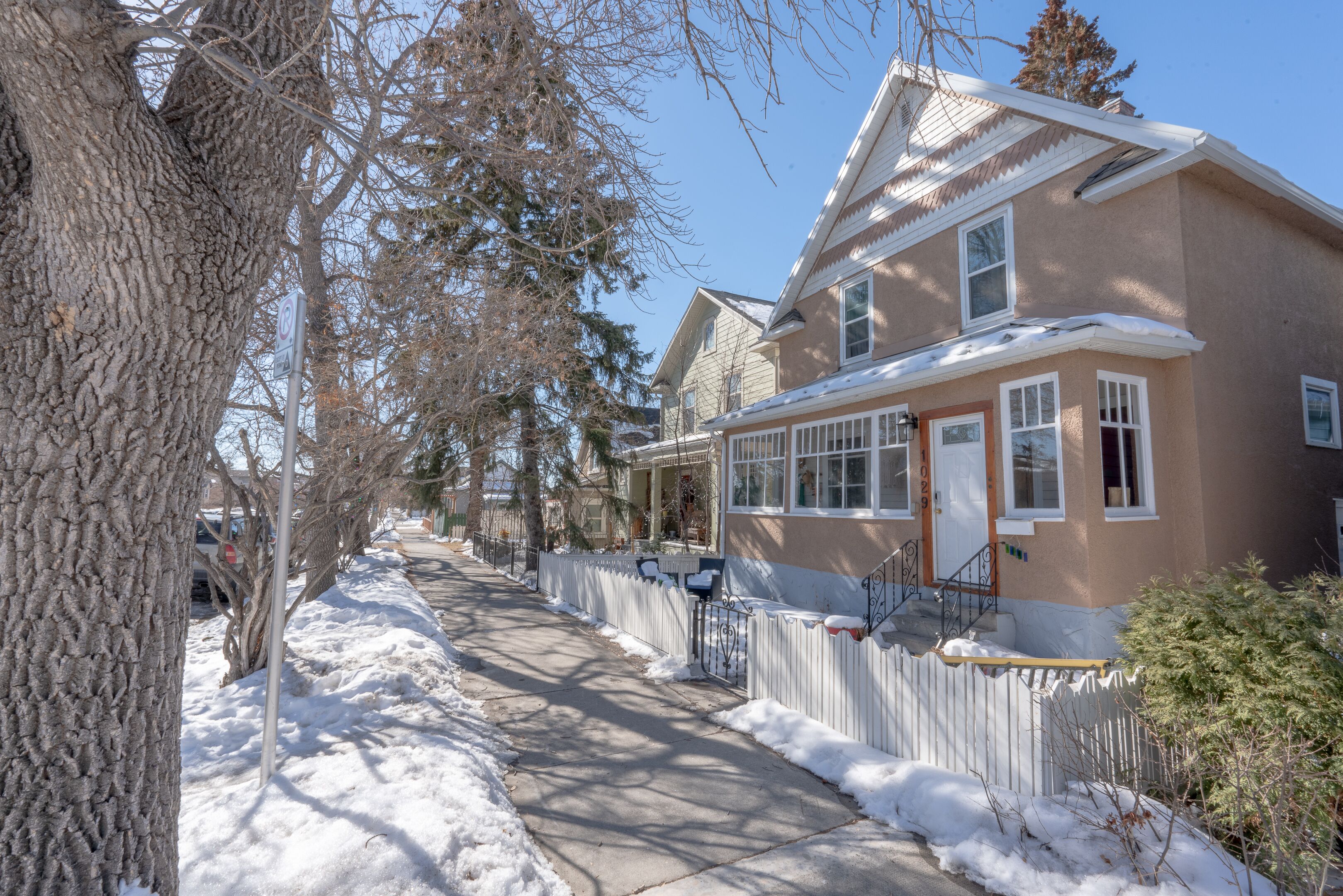 A residential street with houses, a sidewalk, and a snow-covered ground.