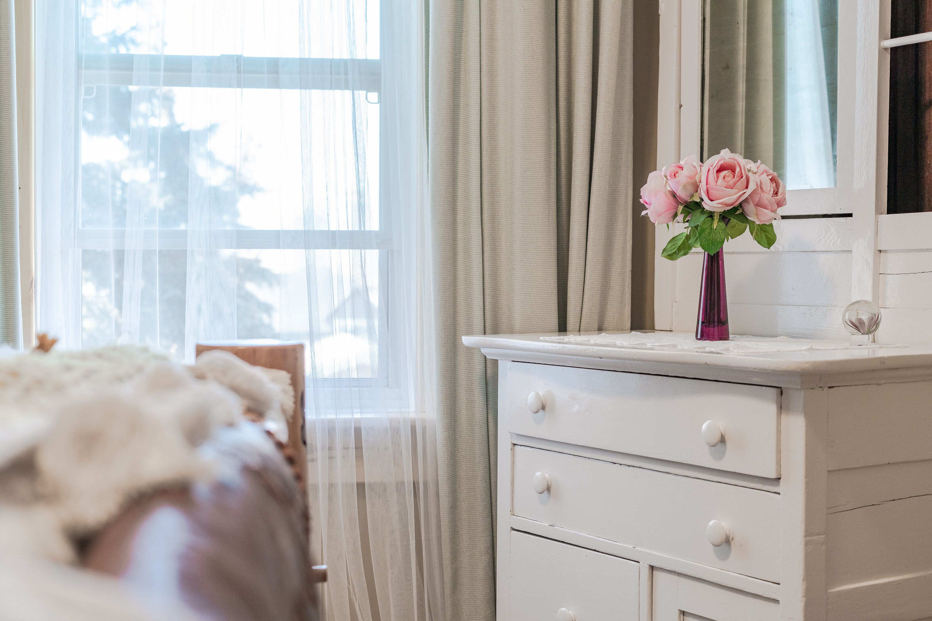 Une chambre à coucher avec un lit, une commode, un vase de fleurs et une fenêtre avec des rideaux.