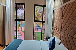 Habitación de hotel con cama, ventanas amplias y vistas a un jardín y edificios.