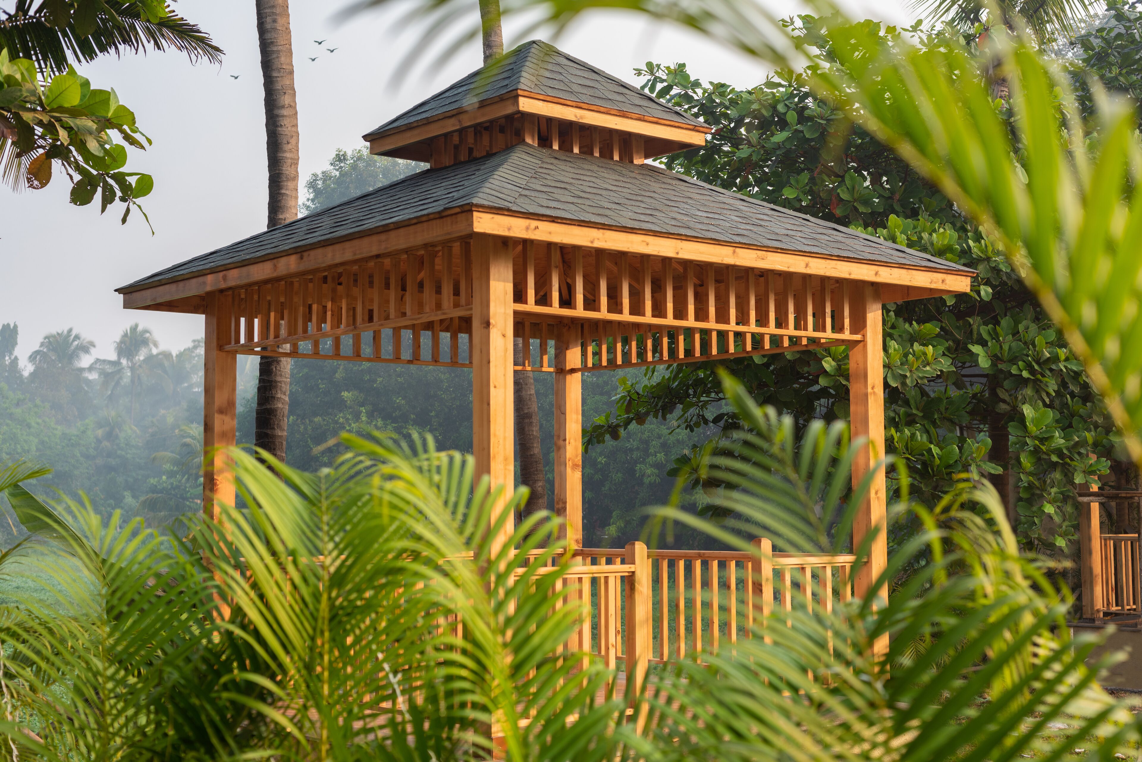A wooden gazebo with a sloped roof, surrounded by lush greenery and palm trees.