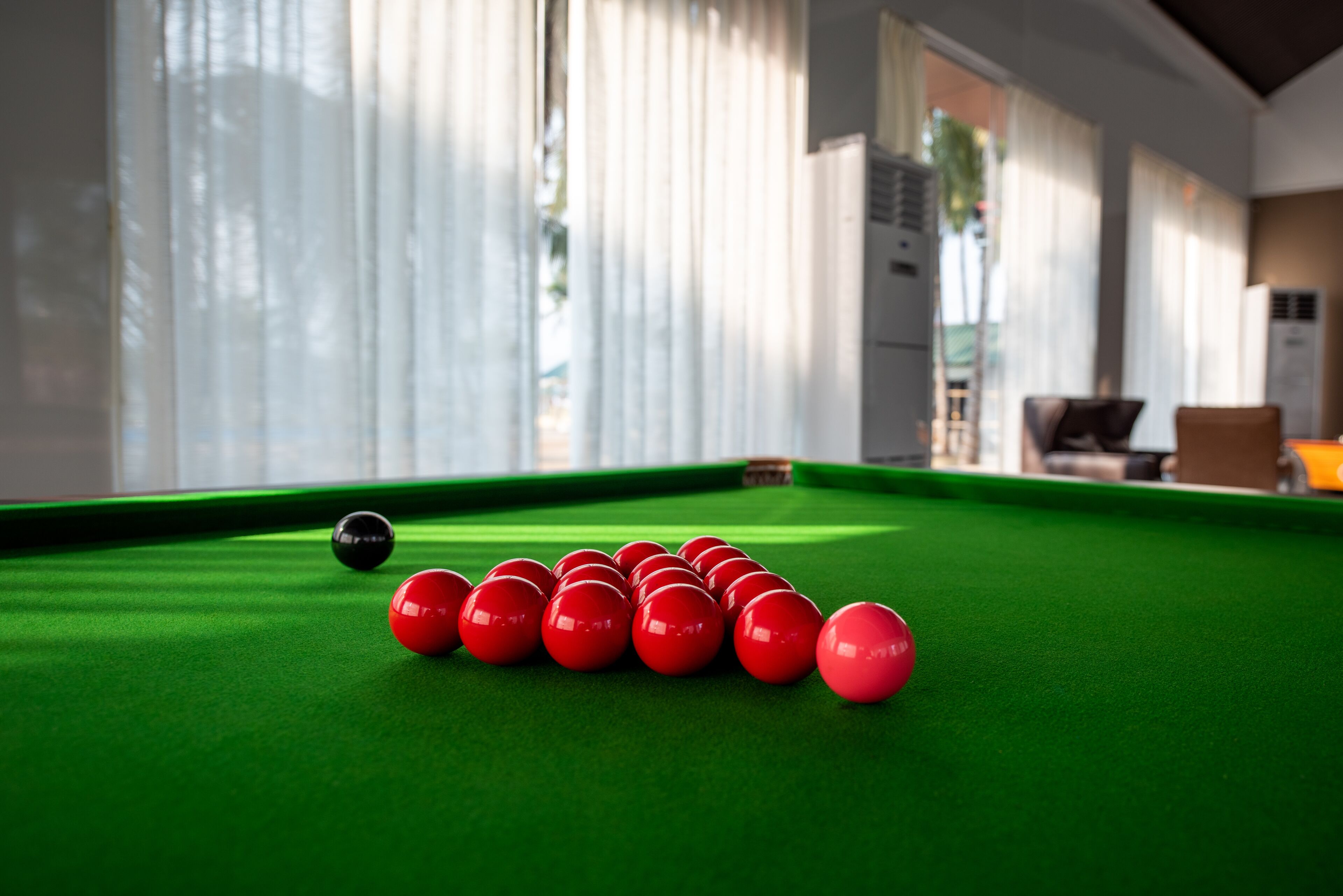 A pool table with red and black balls, a green felt surface, and a room with large windows and curtains.