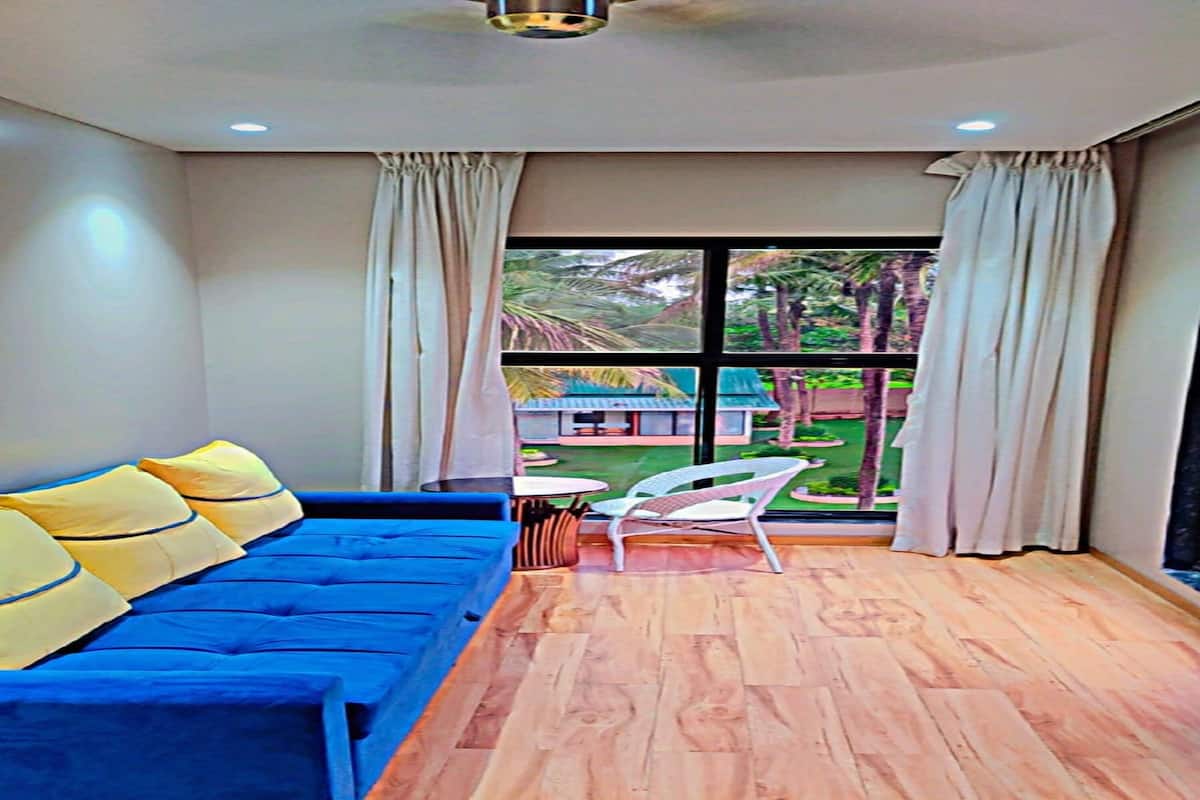 Une pièce avec un canapé bleu, une petite table et une chaise. Il y a une fenêtre avec des rideaux, et de là, on peut voir la verdure à l’extérieur.