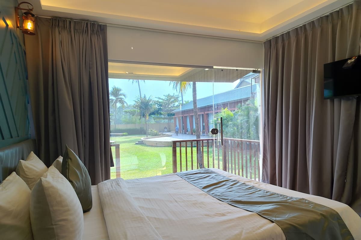 Une chambre d’hôtel avec un lit, des rideaux, une télévision et un balcon donnant sur un immeuble et des palmiers.