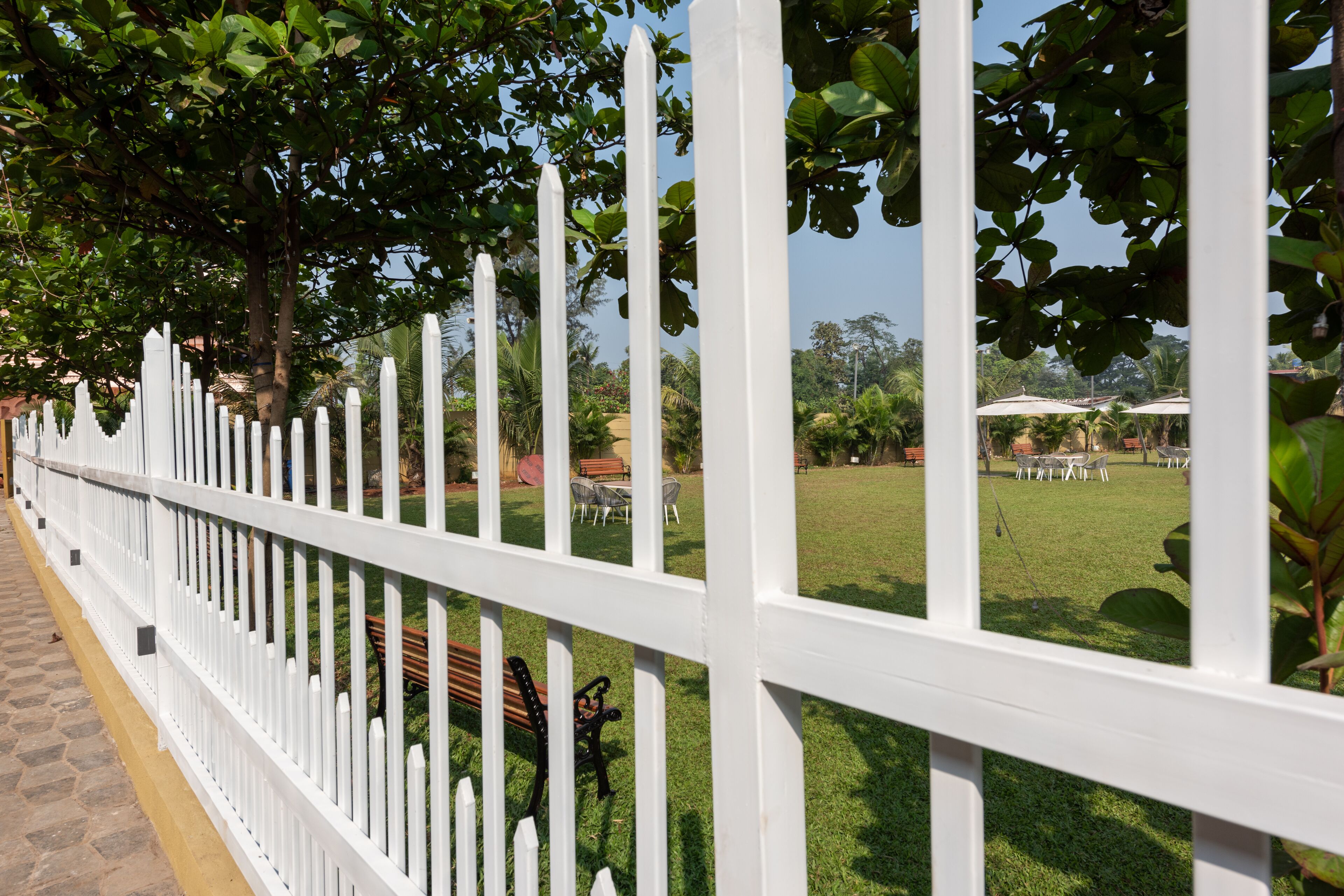 Una cerca blanca con listones verticales, un sendero pavimentado y un área con césped y mobiliario de exterior.