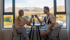 Deux personnes sont assises autour d’une table ronde, devant une bouteille et des verres, et elles ont une vue sur un paysage montagneux grâce à de grandes fenêtres.