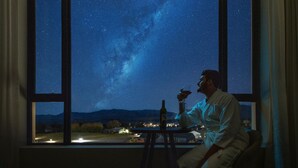Une personne vêtue d’une robe, debout près d’une fenêtre, avec une vue sur un ciel étoilé.