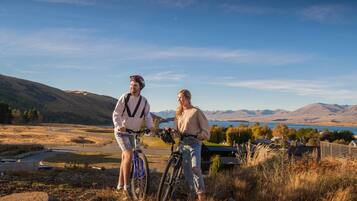Two people standing on a grassy hill with bicycles, overlooking a valley with mountains and a lake.