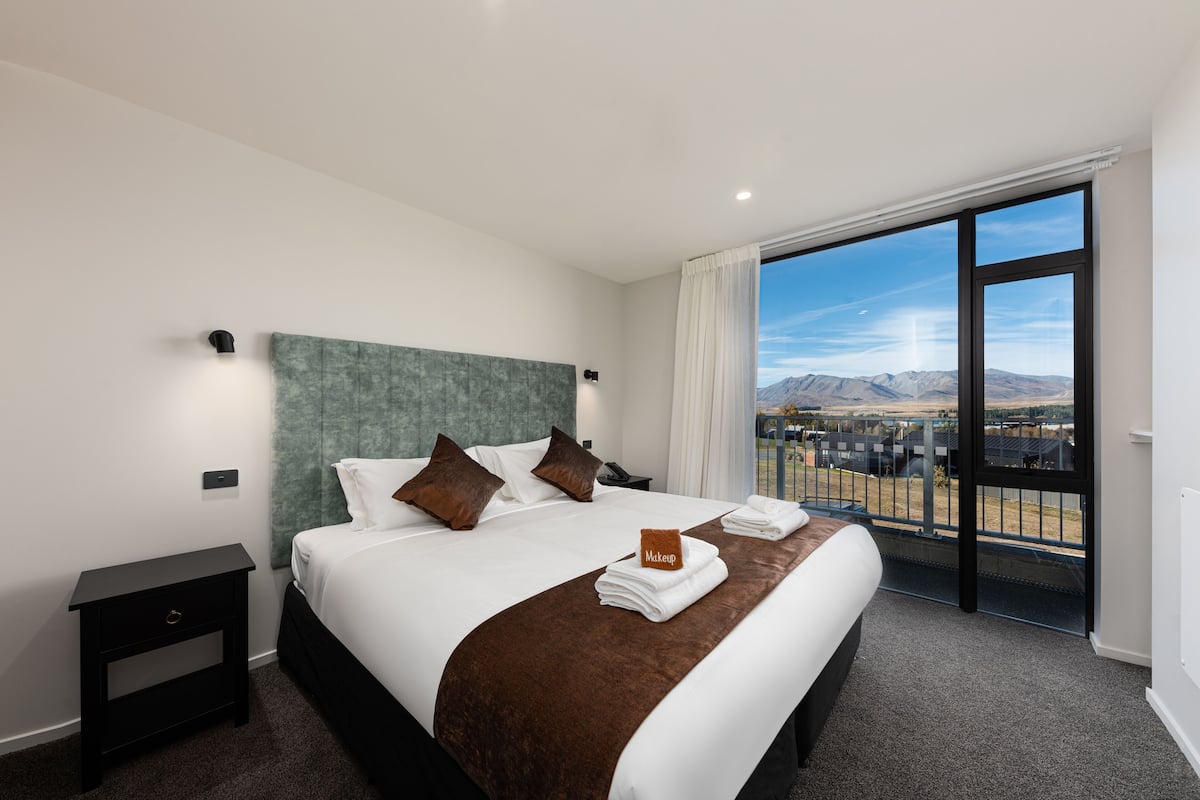 Habitación de hotel con una cama grande, una mesita de noche, vistas a montañas a través de un balcón y un letrero que indica una zona de maquillaje.