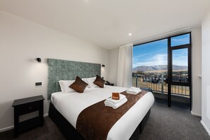 Habitació d'hotel amb un llit gran, una tauleta de nit, vista a les muntanyes a través d'un balcó i un senyal que indica un espai per maquillar-se.