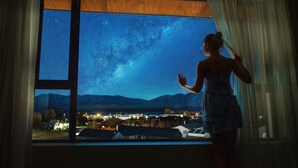 Une personne se tient près d’une fenêtre, d’où l’on peut admirer le ciel nocturne, avec ses étoiles et une chaîne de montagnes visible.