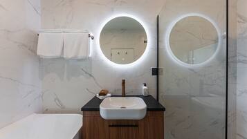 Un baño moderno con pared de azulejos de mármol, un lavabo blanco y dos espejos redondos con iluminación integrada.