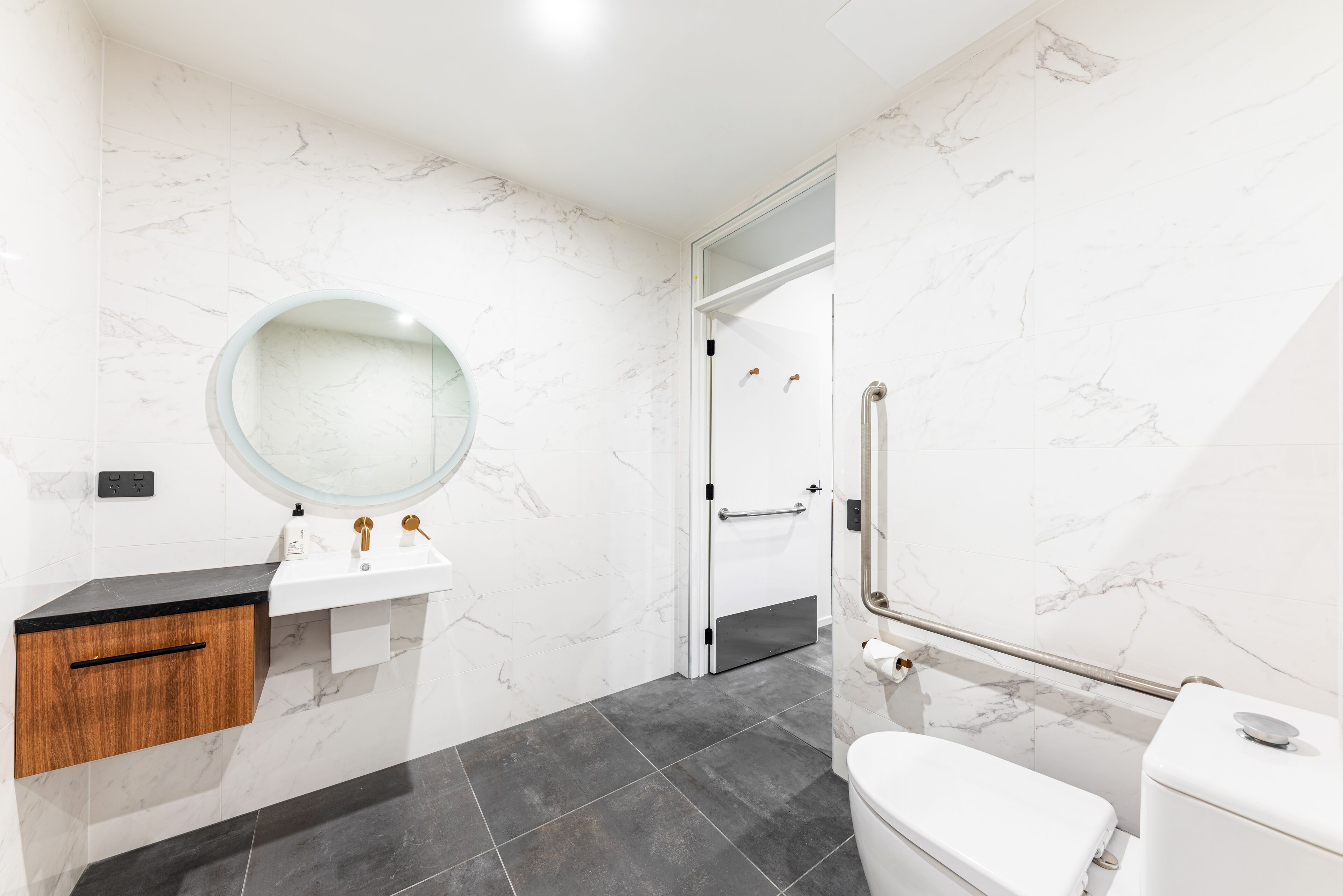 A modern bathroom with a marble wall, a round mirror, a white sink, a wooden cabinet, a toilet, and a shower area.