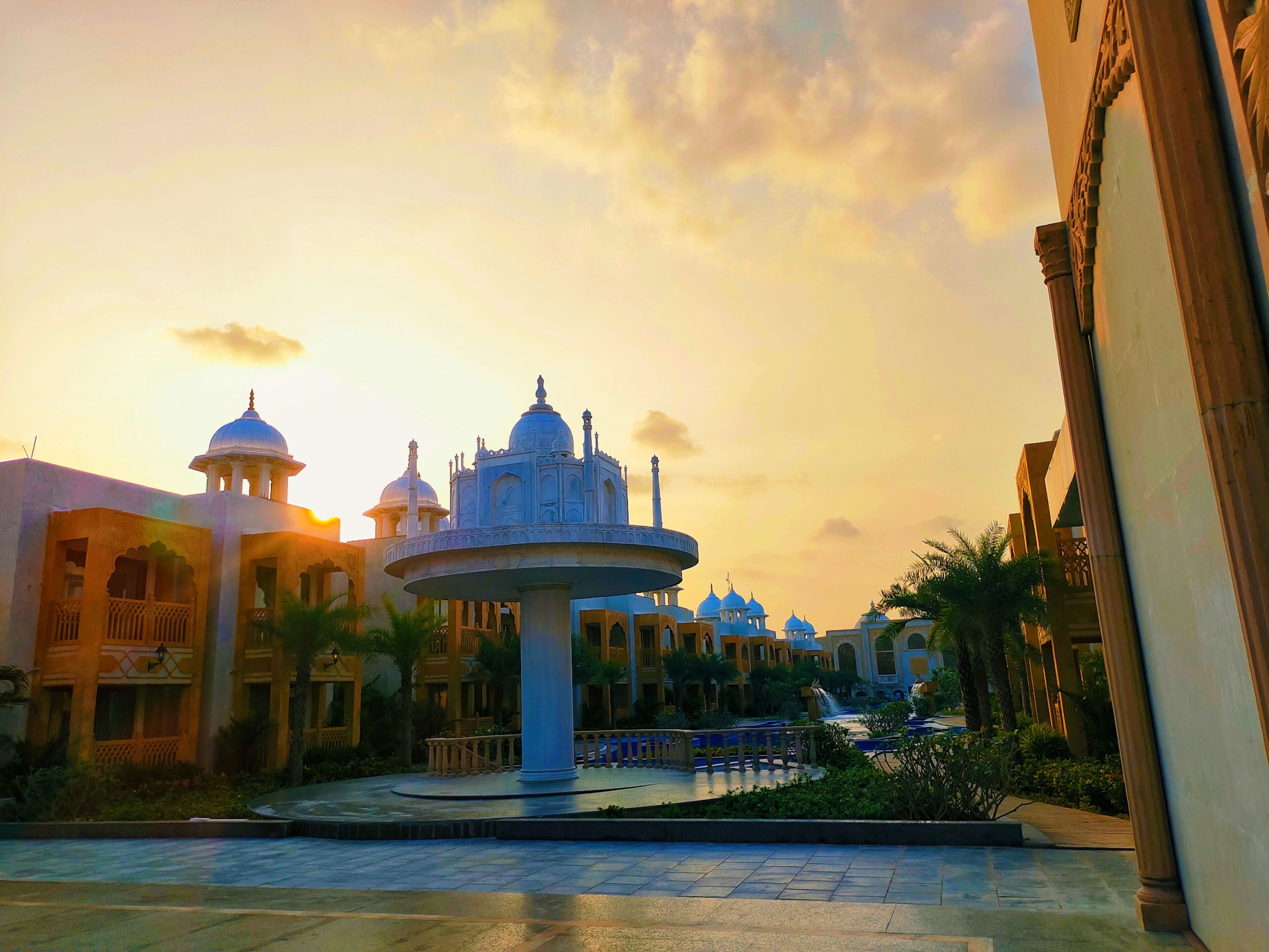 A courtyard with a central fountain, surrounded by classical architecture with domes and arches, under a sunset sky.