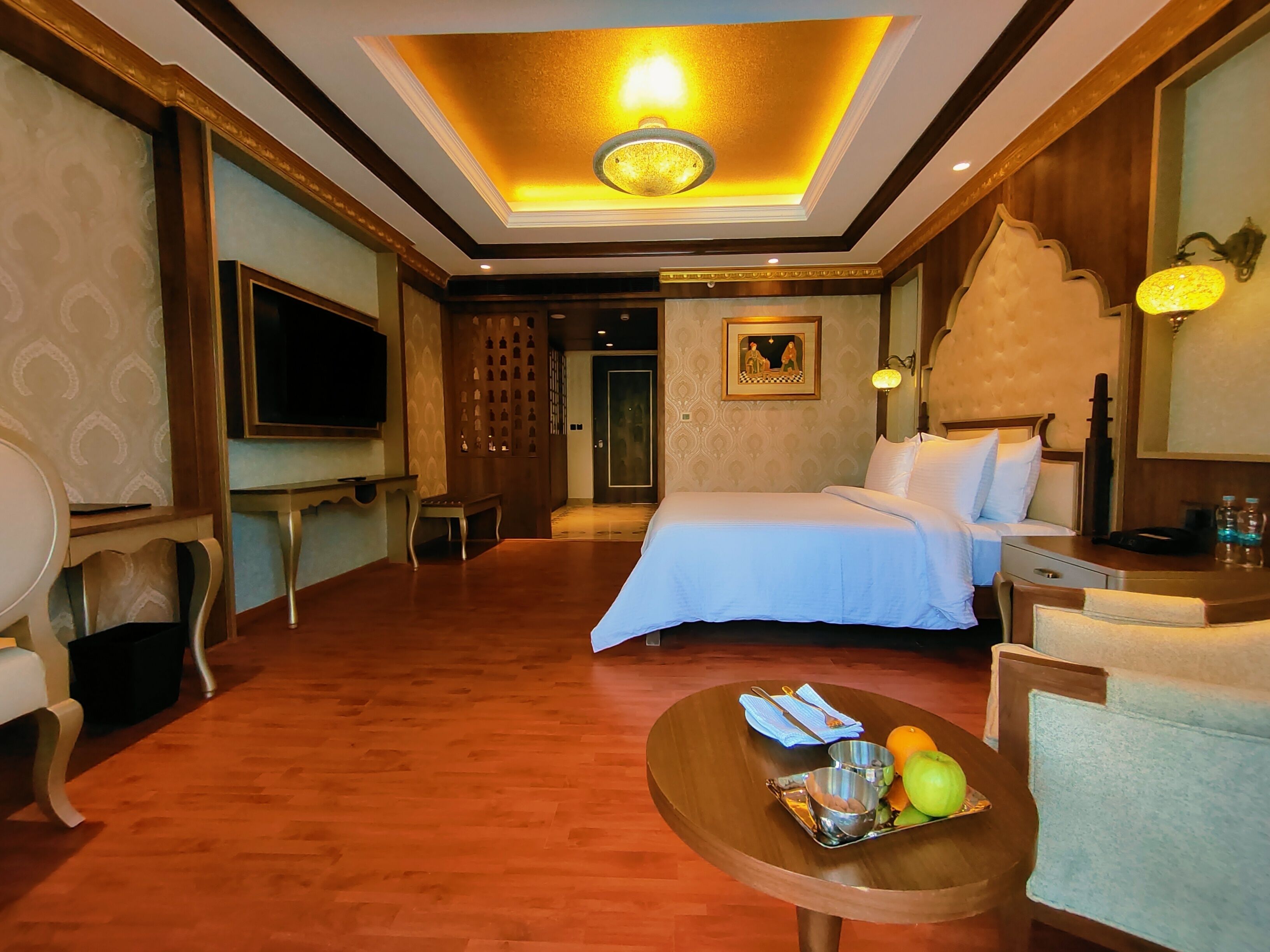A hotel room with a bed, a desk, a chair, a television, and a dining table with fruits and utensils.