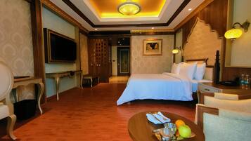 A hotel room with a bed, a desk, a chair, a television, and a dining table with fruits and utensils.