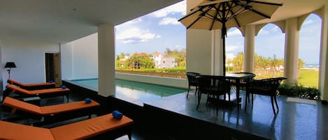 Un área de piscina interior moderna con sillones de descanso y una mesa bajo un gran paraguas.