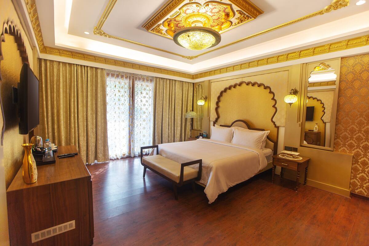 A spacious hotel room with a large bed, a desk with a flat-screen TV, a bedside table, and a large mirror.
