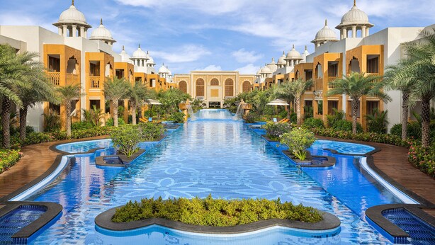 Un área de piscina con varias piscinas, palmeras y un edificio con estructuras abovedadas.