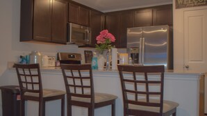 A kitchen with dark wooden cabinets, a stainless steel refrigerator, a microwave, and a sink. There are three wooden chairs with cushioned seats. A vase with pink flowers is on the counter. - Dreamville (Maxwell)