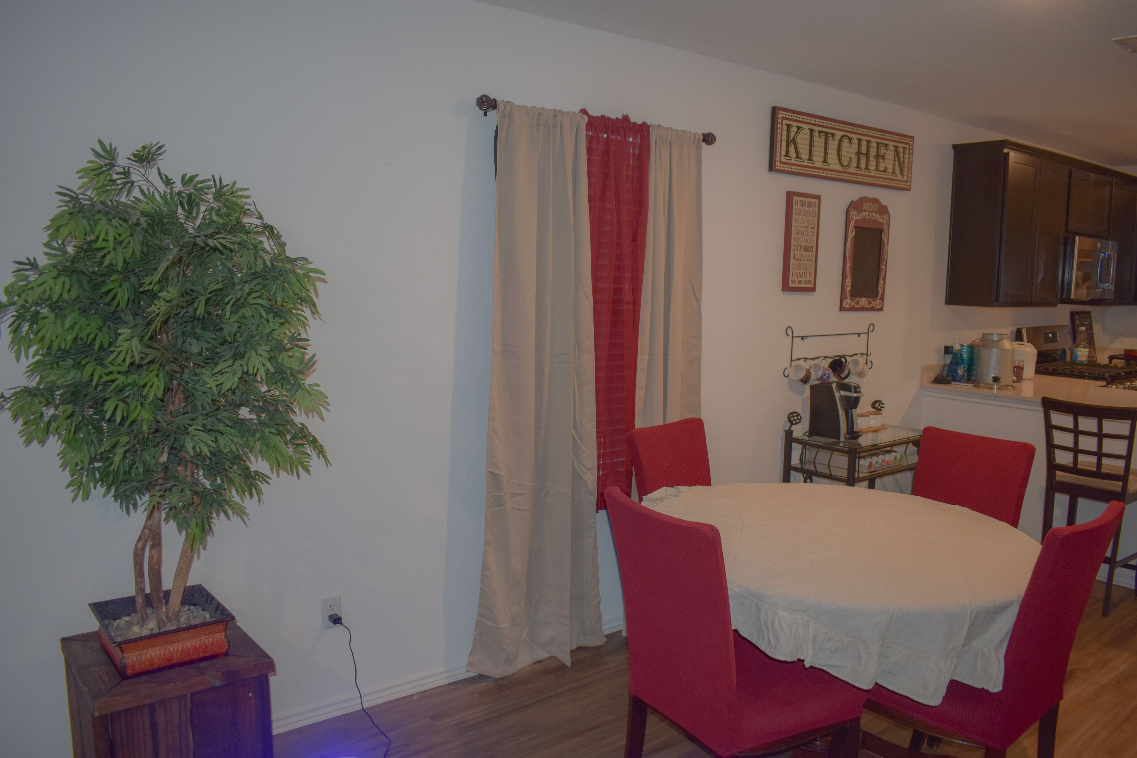A dining area with a round table, red chairs, a potted plant, and a kitchen sign on the wall.
