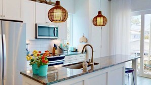 A modern kitchen with white cabinets, a stainless steel refrigerator, a microwave, a sink with a faucet, and two hanging wicker pendant lights. - Modern Duplex, Fenced Yard, Dogs OK, Easy Walk to Beach + Greenway (Carolina Beach)
