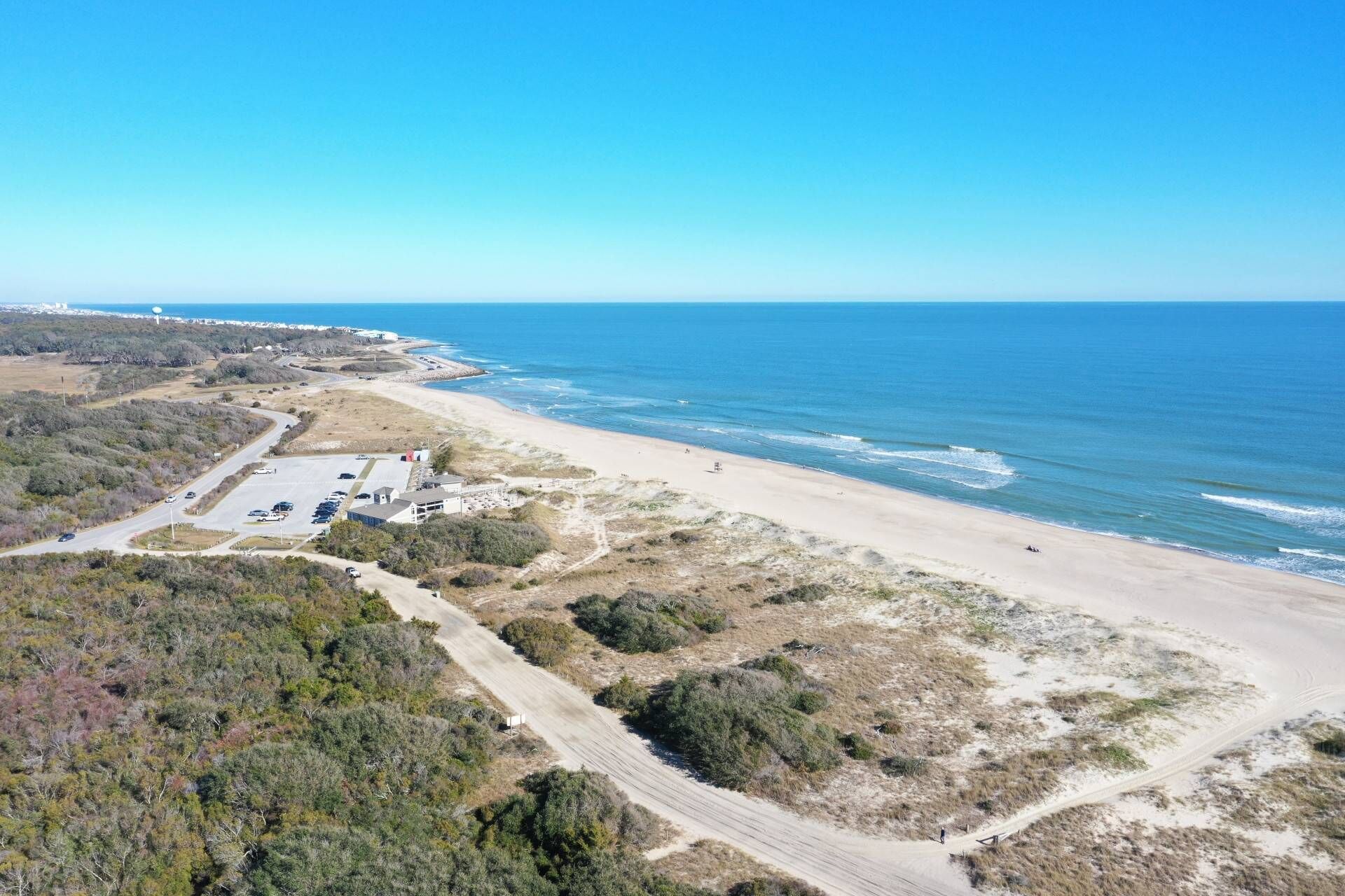 Vista aérea de una zona costera con playa de arena, un estacionamiento y vegetación escasa.