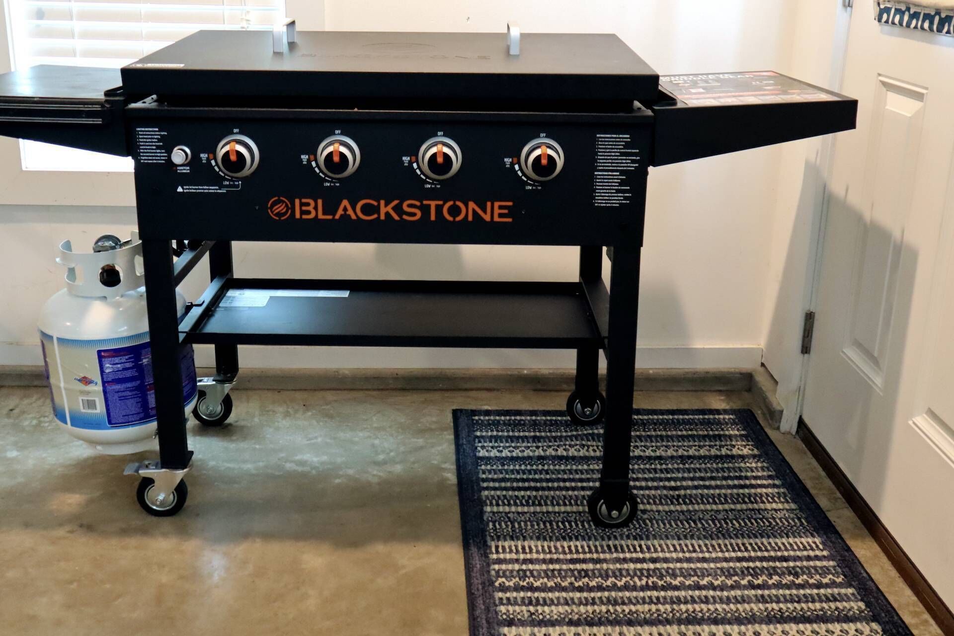 A BLACKSTONE portable grill with four control knobs, a gas tank, and a wheeled base, placed on a patterned rug in a kitchen.