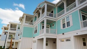 A row of two-story townhouses with white exteriors and teal accents, featuring balconies and garage doors. - Stylish Luxury Townhome w/ Pool, Elevator, 3 Kings & an Ocean View Near the Beach (Kure Beach)
