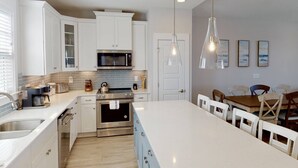 A modern kitchen with white cabinetry, a built-in oven, a microwave, and a large island with a sink. There are pendant lights above the island and a dining area with a wooden table and chairs. - Stylish Luxury Townhome w/ Pool, Elevator, 3 Kings & an Ocean View Near the Beach (Kure Beach)