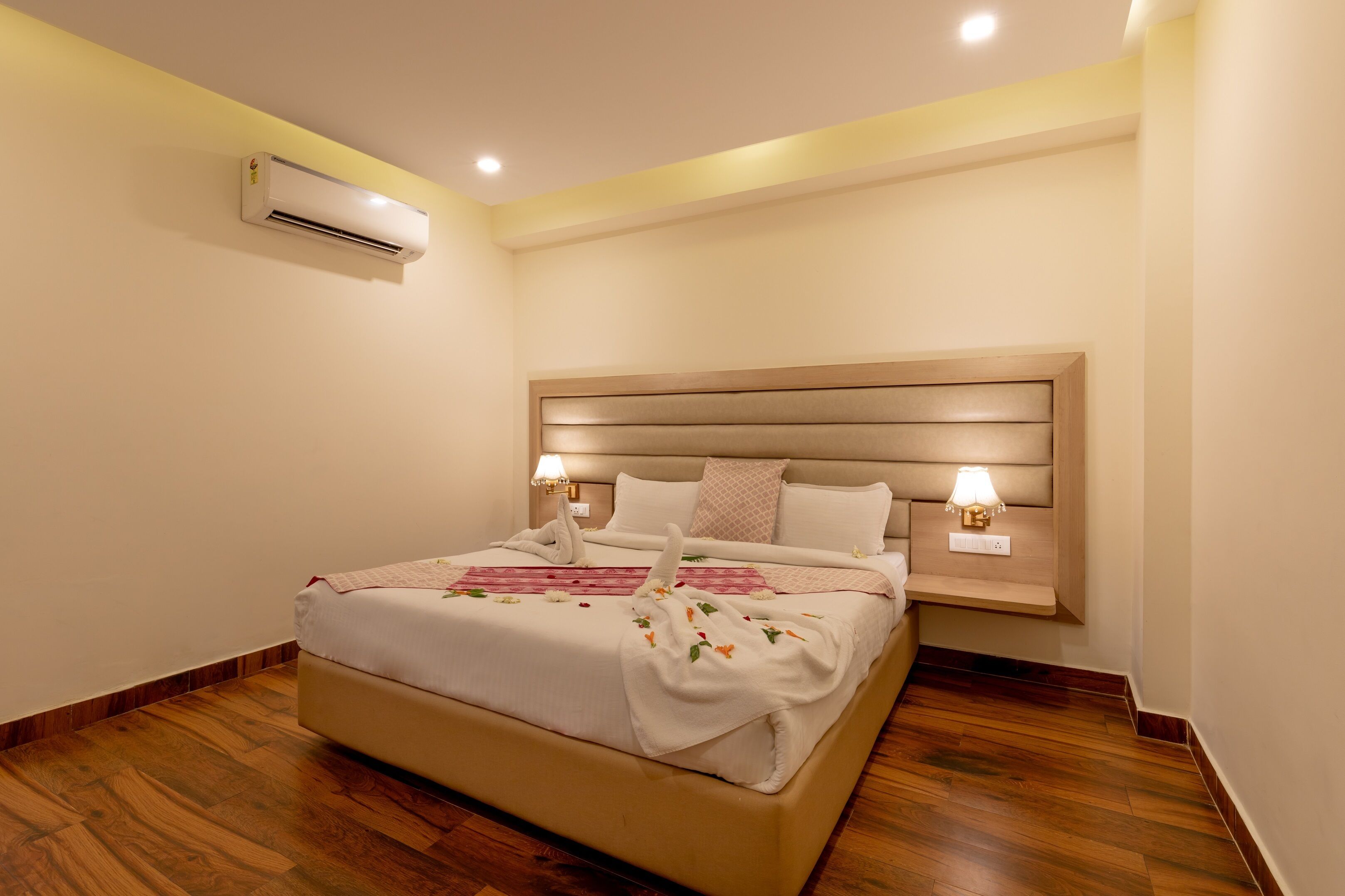 A hotel room with a large bed, bedside lamps, an air conditioning unit, and a wooden floor.