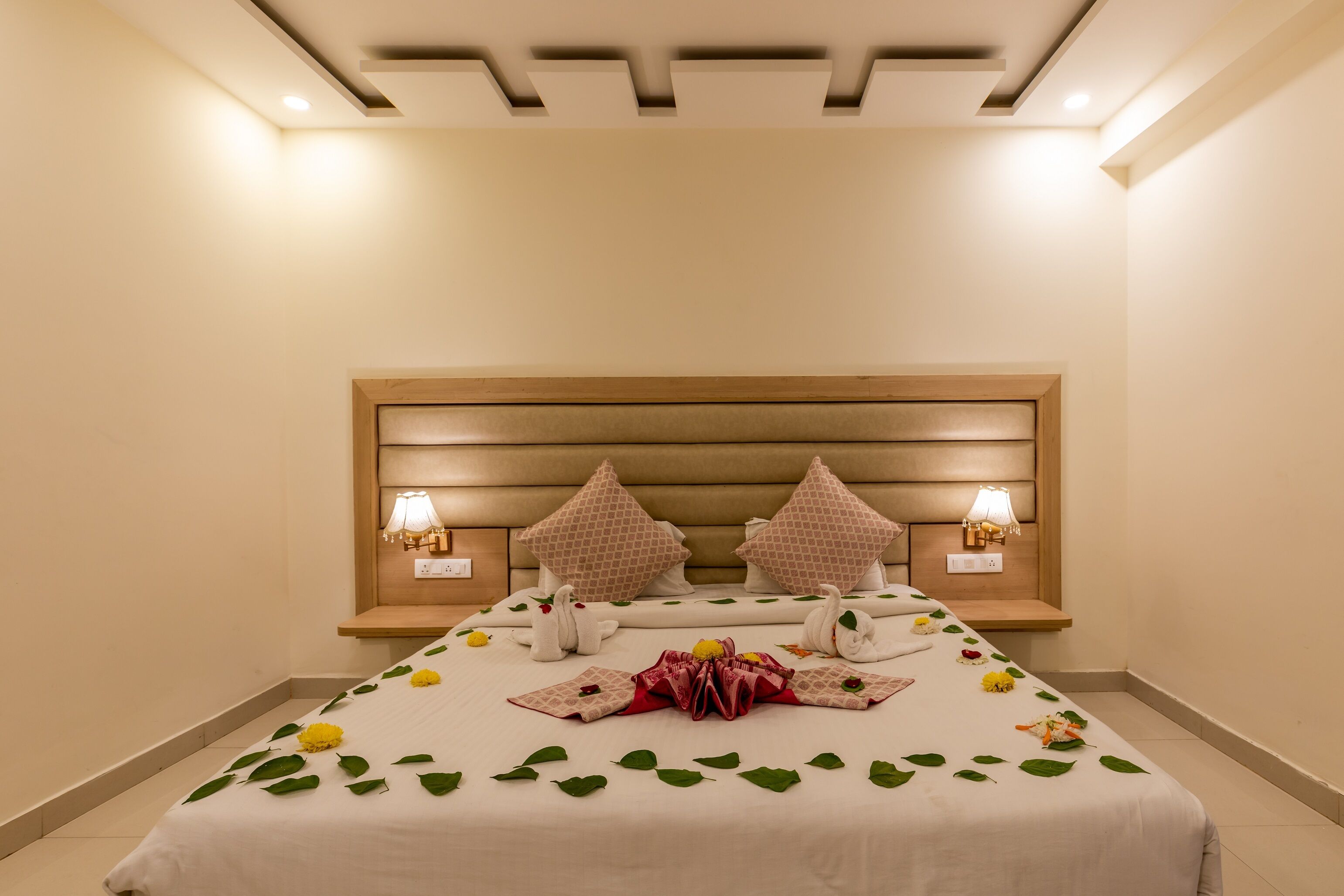 A hotel room with a large bed, two bedside lamps, a wooden headboard, and a decorative arrangement of flowers and leaves on the bed.