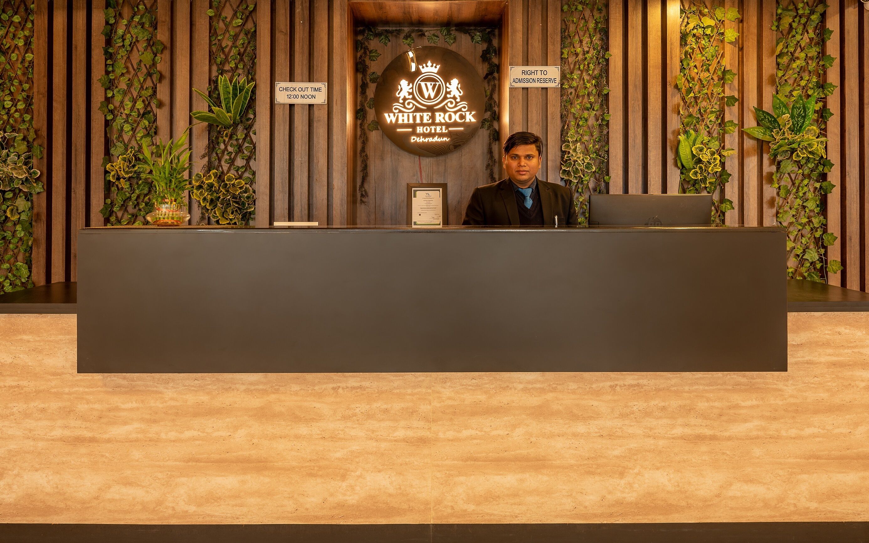 Eine Rezeption im White Rock Hotel in Dehradun mit Hinweisschild für die Auscheckzeit, Schild mit Zugangsbeschränkung und einem Logo mit einer Krone und Löwen.