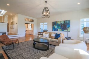 A spacious living room with a modern chandelier, a large abstract painting, and a view of the kitchen. - Ocean Views, Two Master Suites, Screened Porch & Access to Ocean Point Community Pool & Cabana (Isle of Palms)