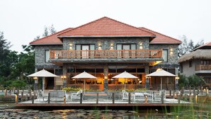 Een gebouw met twee verdiepingen, voorzien van een rood pannendak, terras met parasols en een vijver met waterlelies.