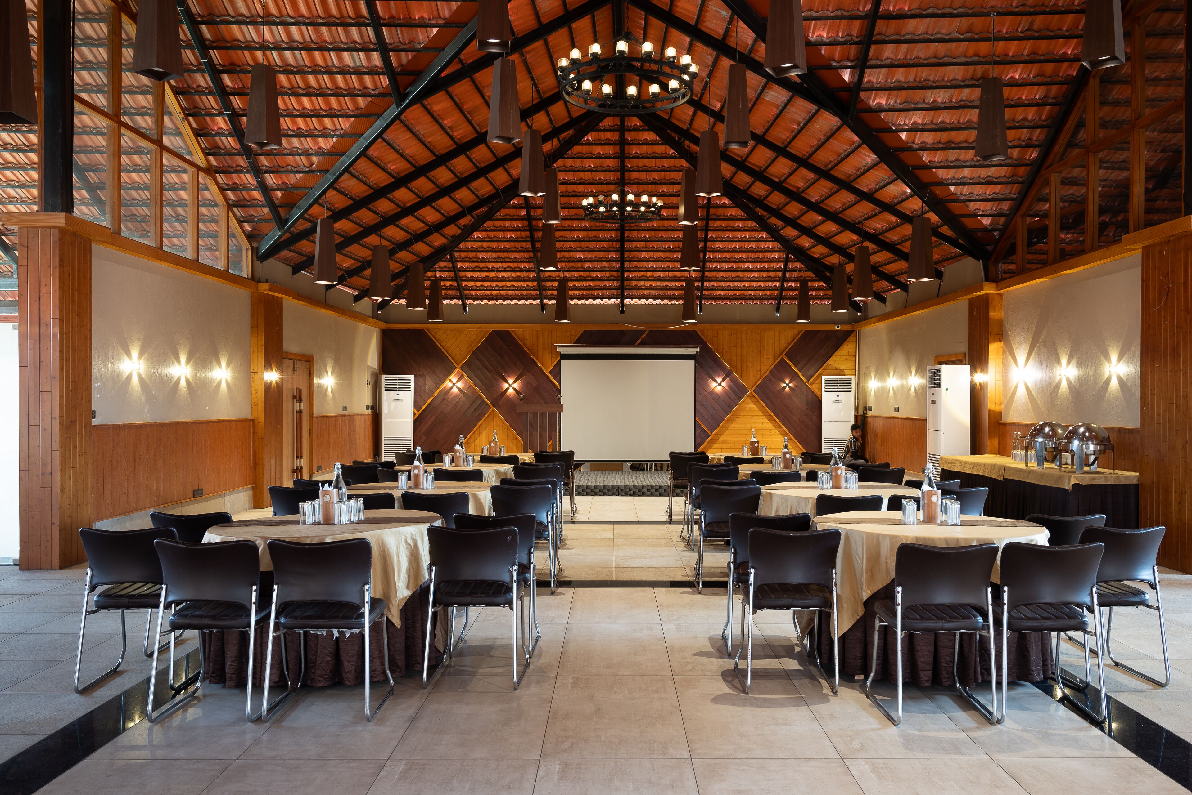 Una espaciosa sala de conferencias con varias mesas y sillas, un techo alto con una estructura de madera, y una pantalla grande al frente.