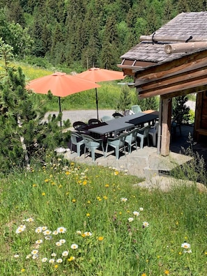 Un espace de restauration extérieur rustique, avec une table et des chaises, entouré d’un paysage naturel agrémenté d’arbres et de fleurs.