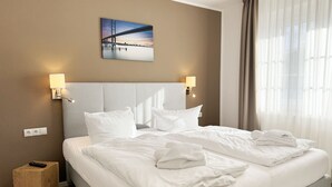 A hotel room with a large bed, two bedside lamps, a painting of a bridge, and a view of a body of water. - House B - Whg 01 with terrace - F: Lotsenberg Apartments with 8 apartments (Mönchgut)