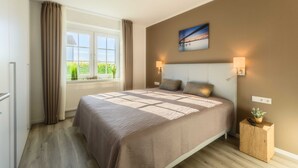 Quarto com uma cama grande, janela com cortinas, um quadro na parede e uma pequena planta sobre uma mesa de cabeceira.