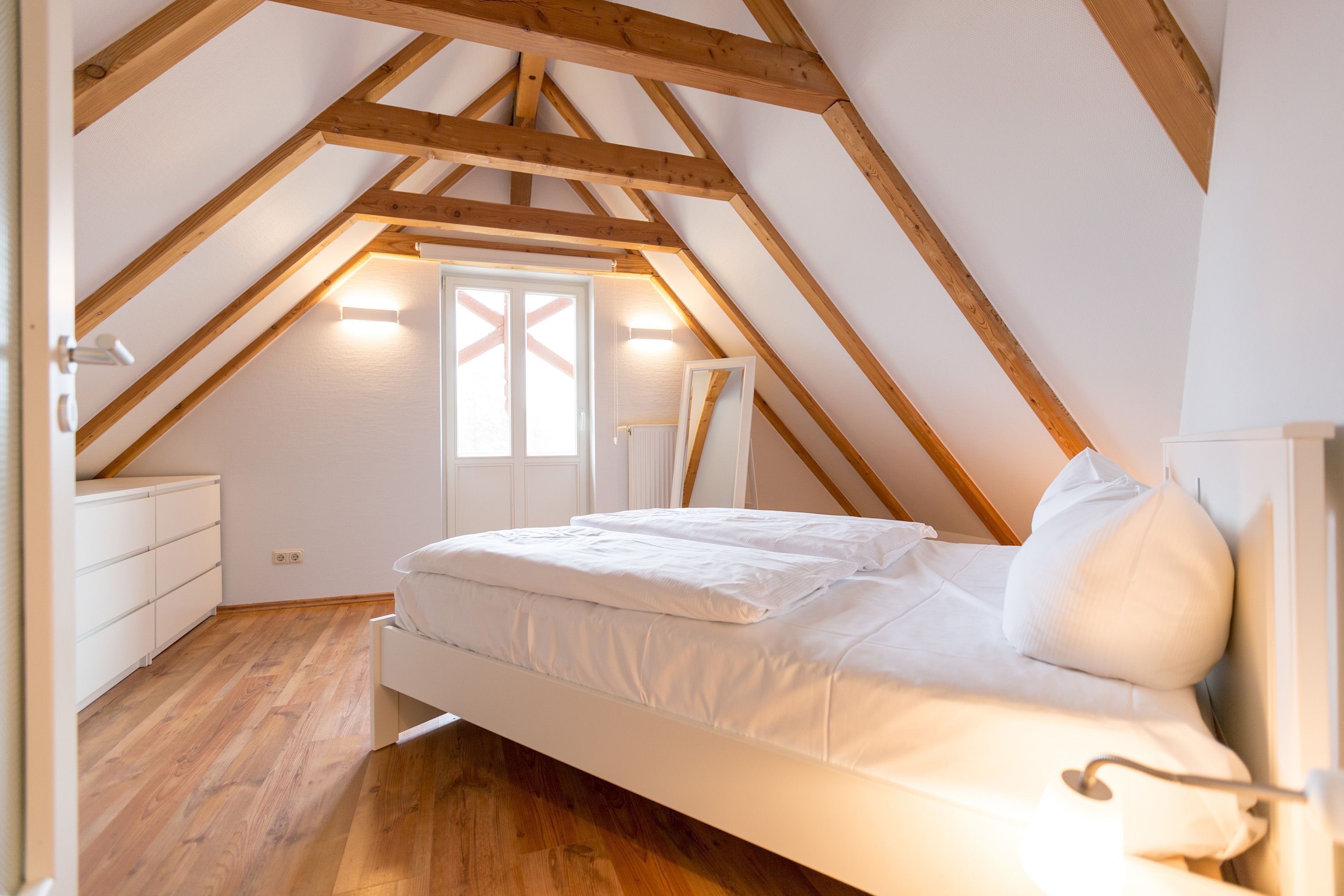 Un dormitorio con techo inclinado, vigas de madera, una cama con ropa blanca y un ventanal con vistas.