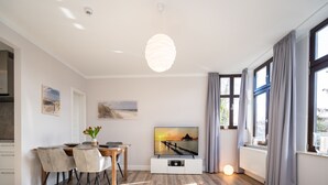 Ein modernes Hotelzimmer mit Holzboden, einer Essecke mit Tisch und Stühlen, einem Fernseher und großen Fenstern mit Vorhängen.