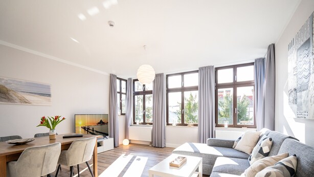 Ein modernes Wohnzimmer mit Sofa, Couchtisch und Essbereich. Große Fenster mit Vorhängen, ein Gemälde an der Wand und Blick ins Freie.