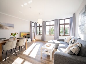 Una sala moderna con sofá, mesa de centro y comedor. Hay ventanas grandes con cortinas, un cuadro en la pared y vistas al exterior.