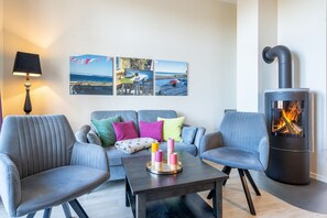 Ein modernes Wohnzimmer mit einer Couch, zwei Sesseln, einem Couchtisch mit Kerzen, einem wandmontierten Kamin und einer Lampe.