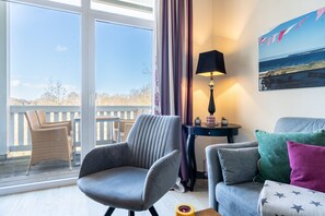 Een kamer met uitzicht op zee, een rieten stoel, een grijze fauteuil, een grijze bank en een lamp op een bijzettafeltje.