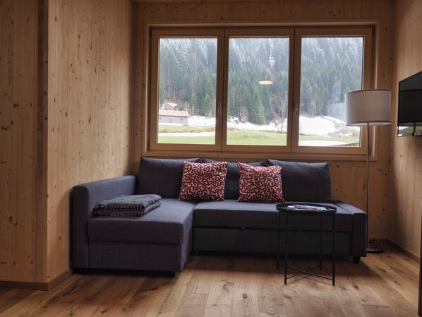 A wooden-paneled room with a sectional sofa, a small table, and a window with a view of a forest and a building.