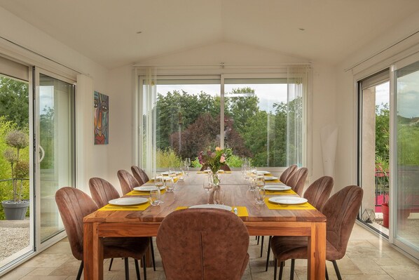 Uma sala de jantar com uma mesa grande posta para refeição, rodeada por cadeiras, grandes janelas e vista para o verde lá fora.