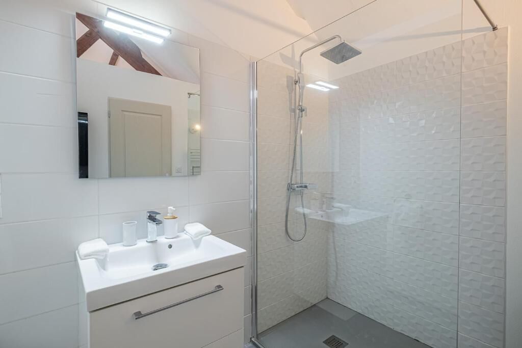 Une salle de bain moderne avec une douche à parois de verre, un lavabo blanc avec un miroir au-dessus, et un mur recouvert de carreaux blancs.