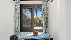Une pièce avec une table, des chaises, une fenêtre avec des rideaux et un saladier de fruits sur la table.
